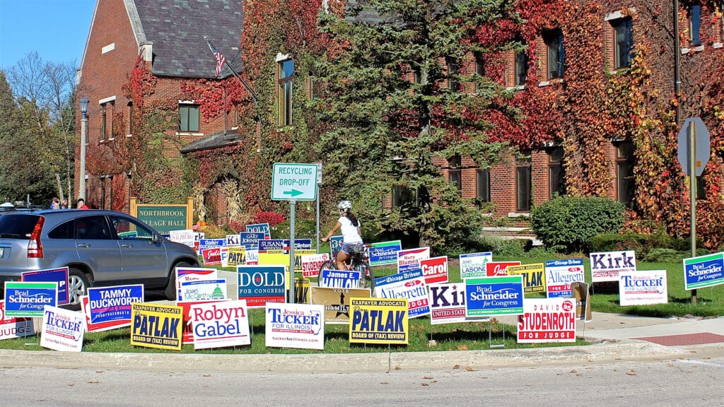 Northbrook, IL, USA - November 6, 2016: Biker riding on sidewalk through election campaign political lawn signs for local, state and national candidates outside polling place during early voting at Village Hall.