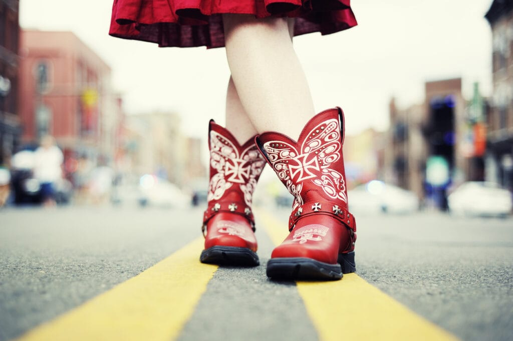 A cowgirl in red cowboy boots and dress has her legs crossed, standing on the double yellow line in the middle of a city street in Nashville. 