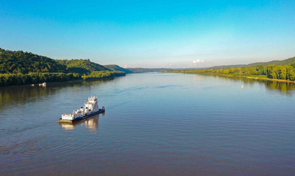 An aerial view captures a tugboat navigating the Ohio River between Indiana and Kentucky, gracefully sailing on a river amidst mountainous terrain.