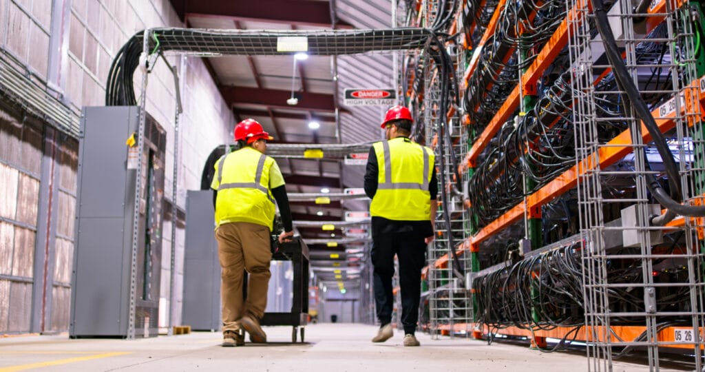 Two technicians walking past racks of equipment in a data center for cryptocurrency mining, cloud services and AI computing in a large, temperature controlled warehouse in a remote location in  Stutsman County, North Dakota.